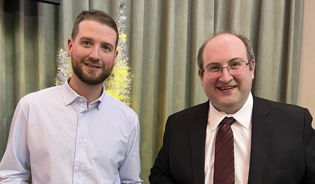 Jeune homme professionnel en costume et épaules décontractées lors d'un événement d'entreprise, debout près d'un arbre décoratif, dans un cadre intérieur chaleureux.