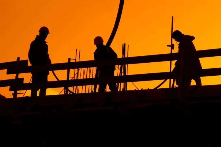 Construction workers silhouetted against an orange sky at sunset, wearing hard hats on a building site.
