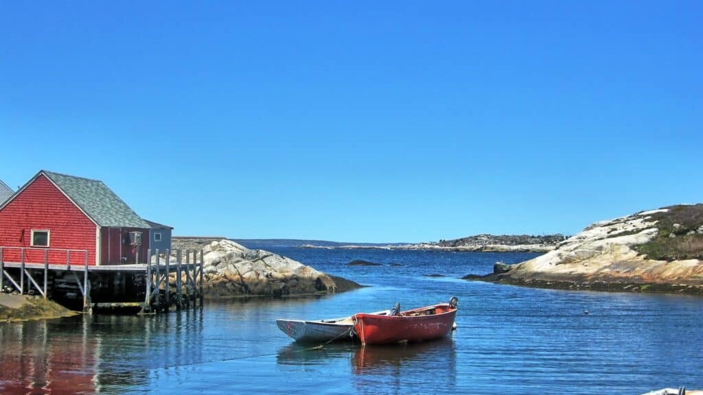 Bateau rouge amarré dans un petit port de pêche avec maison en bois rouge, paysage côtier au Québec, ambiance paisible, vue sur l'eau et les rochers naturels.